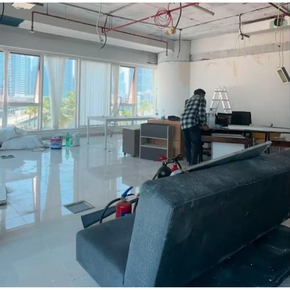 Interior office renovation in progress, featuring a worker at a desk, modern furniture elements, large windows with city views, and construction materials like paint and tools scattered around.